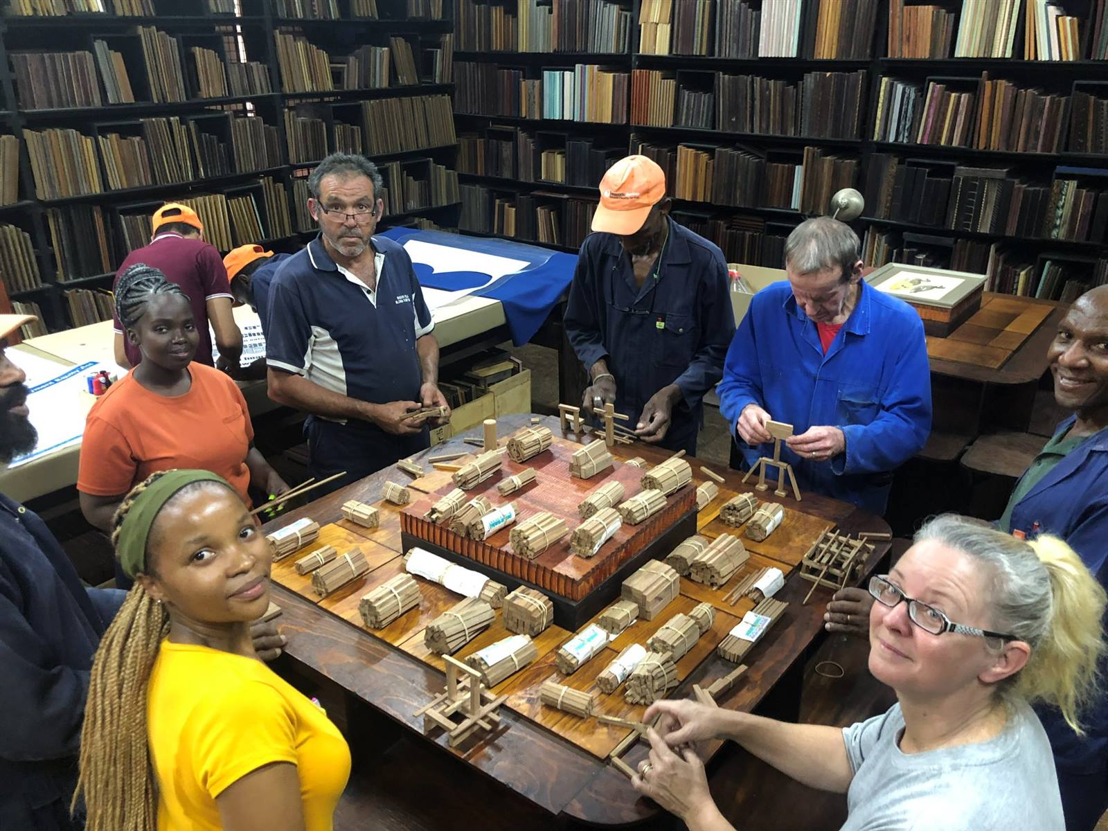 The team and visitors round a table of bundled sticks, building small frames in the Woza Woza Workshop